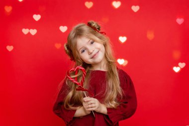 Portrait of a cheerful dreamy little girl with a heart shaped candy in her hands isolated on a red background