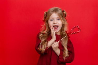 Portrait of a cheerful dreamy little girl with a heart shaped candy in her hands isolated on a red background