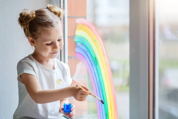 Uma menina com cabelo loiro desenha um arco-íris no vidro com tintas . — Fotografia de Stock