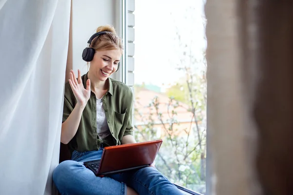 Una mujer rubia feliz usando una computadora portátil para videollamadas a amigos y padres, una chica sentada en casa en la ventana . — Foto de Stock