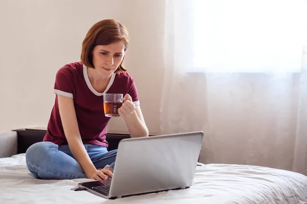 Young red-haired woman on the bed with a laptop. The girl reads the bad news, is upset.