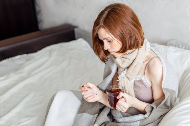 Yatağında atkıyla oturan hasta bir genç kadın boğaz ağrısı çekiyor. Sıcak çay içiyorum. Ateşli bir kadın battaniyeye sarılmış..