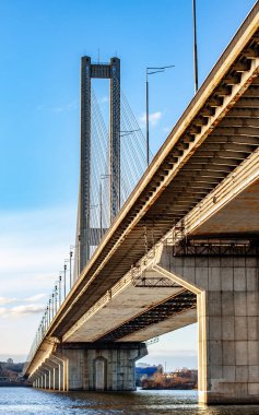 Bu çarpıcı görüntü, aşağıdan bakıldığında modern bir asma köprü sergiliyor. Mimari zarafetini parlak mavi gökyüzüne ve sakin sulara vurguluyor..