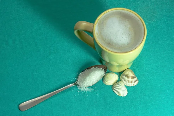 A cup of cappuccino with a spoon of sugar and seashells on a turquoise background