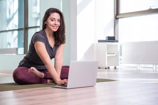 Pandemia sırasında dizüstü bilgisayarla yoga yapan genç bir kadının fotoğrafı. Güzel kız sınıfta yoga yapıyor. Yoga stüdyosu eğitmeni. Bulanık arkaplan