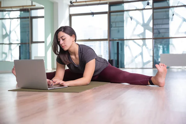 Pandemia sırasında dizüstü bilgisayarla yoga yapan genç bir kadının fotoğrafı. Güzel kız sınıfta yoga yapıyor. Yoga stüdyosu eğitmeni. Bulanık arkaplan