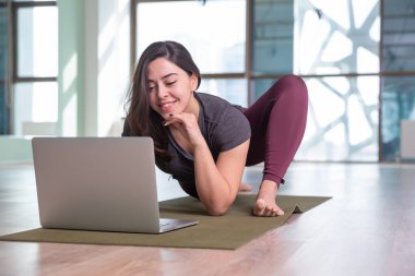 Evde dizüstü bilgisayarla yoga yapan genç bir kadının fotoğrafı. Güzel kız sınıfta yoga yapıyor. Yoga stüdyosu eğitmeni. Bulanık arkaplan