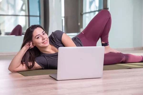 Фото молодої жінки, яка практикує йогу з ноутбуком в приміщенні. Красива дівчина практикує йогу в класі. Інструктор студії йоги. Розмитий фон — стокове фото