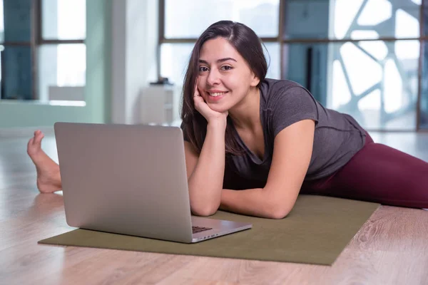 Fénykép fiatal nő gyakorló jóga laptop beltéri. Gyönyörű lány jógázik az osztályban. Jóga stúdió oktató. Homályos háttér — Stock Fotó
