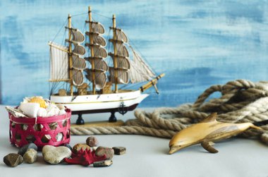 Hatıra botu, halat, ahşap yunus, çakıl taşları, kızıl deniz yıldızı, açık gri arka plan ve mavi arkaplan üzerinde deniz kabukları eşliğinde sepet..