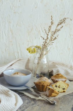 Kahvaltı çörekleri pasta kağıdının üzerinde ve gri-mavi bir kasenin içinde, bir bıçağın yanında, hafif bir havlu ve kuru çiçekler kavanozun içinde, mermer desenli ve açık arkaplanlı bir masaya karşı duruyor..