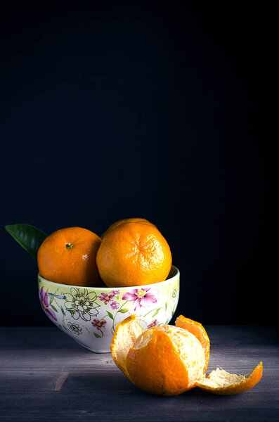 Tangerines lying in a white bowl with a pattern, one lying on a dark table in a peeled peel.