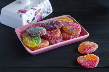 Siyah ahşap bir masanın üzerinde, beyaz bir porselen kapağın yanında, pembe bir tabağın üzerinde duran renkli yuvarlak marmelat..