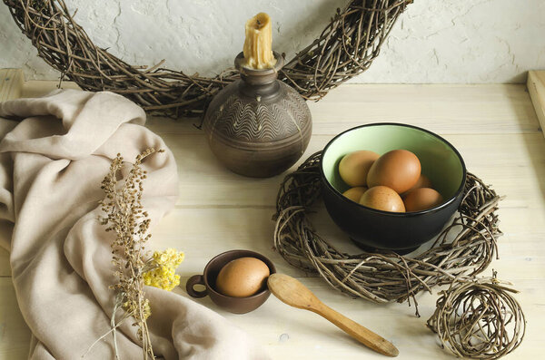 A black-and-green bowl with chicken and homemade eggs and a wicker wreath on a light background, next to a small Cup with an egg and a wooden spoon, a towel and dry flowers, and a clay candle holder with candles. 