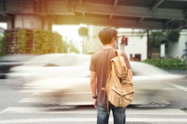 Arka planda bulanık bir arabayla sırt çantasıyla yol kenarında seyahat eden genç bir gezginin defocus 'u. Karşıya geçmeyi bekliyor. Klasik Ton ve Seçici Yumuşak Odak. Seyahat ve seyahat kavramı