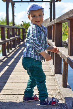 Çocuk parkta ahşap bir köprüde yürüyor, duruyor ve bir şeye bakıyor.