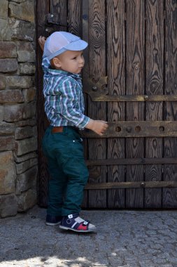 Tişörtlü, yeşil pantolonlu ve şapkalı küçük bir çocuk eski bir mahzene büyük bir ahşap kapı açıyor.