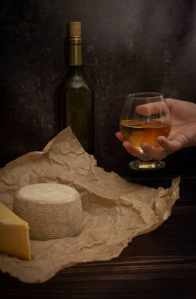 cheese on kraft paper with a bottle of wine and a glass in a male hand on a dark background