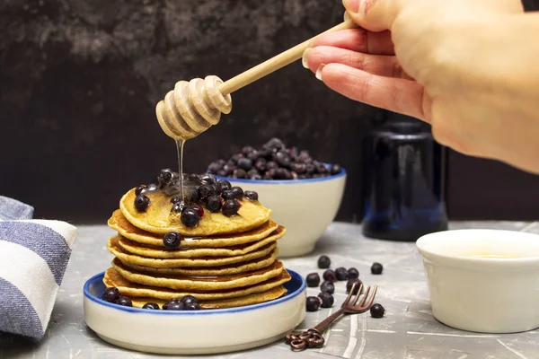 Honey flows from a wooden spoon to pancakes. Wooden spoon for honey in the hand. Pancakes with blueberries and honey on a plate.