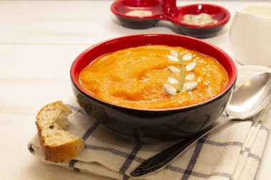 Beyaz ahşap bir masanın üzerinde turuncu kremalı kabak ve havuç çorbası ile birlikte bir tost, kaşık, süt sürahisi ve tohumların yanında. Portakal sebzesinden yapılmış vejetaryen çorbası..
