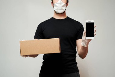 Koruyucu maske takan bir kurye ve tıbbi eldivenler izole edilmiş beyaz bir arka plana karşı bir kutu ve beyaz bir akıllı telefon tutuyor.