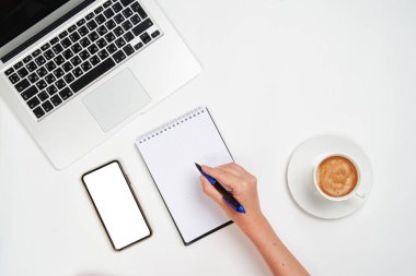 Not defterine kalem, dizüstü bilgisayar, akıllı telefon ve işyerindeki bir fincan kahveyle yazı yazanın üst görünümü