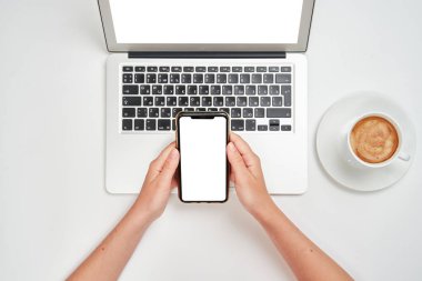 Üst Manzara. Kız akıllı telefonu dizüstü bilgisayarın üstünde tutuyor. Çalışma alanı. İzole edilmiş beyaz bir arkaplanda düz bir zemin