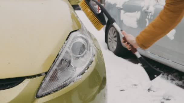 院子里穿着暖烘烘的棕色夹克的女人把车灯从雪中擦干净 — 图库视频影像