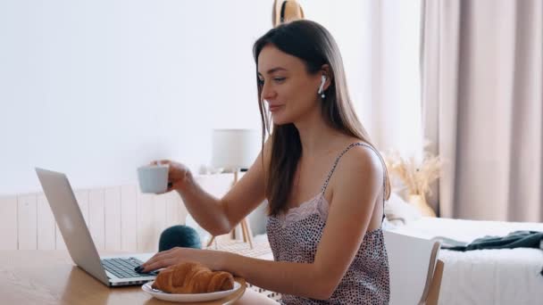 Hermosa chica sonriente feliz en ropa de noche se sienta a la mesa en la mañana soleada escribiendo un mensaje en su computadora portátil — Vídeos de Stock