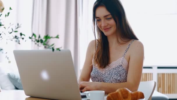 Linda menina sorridente feliz em roupas de noite senta-se à mesa na manhã ensolarada digitando uma mensagem em seu laptop — Vídeo de Stock