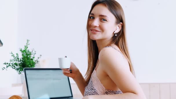 Menina bonita vestida de roupa de noite ouvir música em fones de ouvido sem fio senta-se à mesa e segura caneca de café — Vídeo de Stock