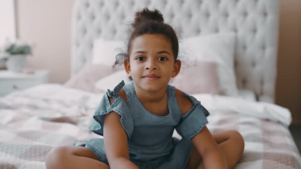 Pretty happy smiling afro american girl child with curly hair sits on bed in cozy bedroom wear blue chintz dress — ストック動画