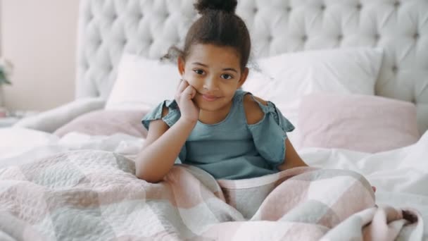 Linda menina negra afro-americana sorridente filha sentar-se em confortável cama de luz com colchão ortopédico no quarto brilhante bebê jovem de bom humor coberto cobertor em estado alegre elevado brincalhão — Vídeo de Stock