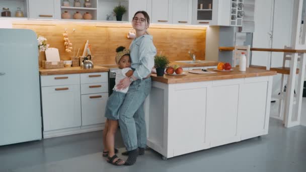 Caring attractive young woman mother and african american girl child happy daughter hug in bright kitchen with fridge dishes stove for cooking fruit on table concept of happy family in apartment — Stock Video