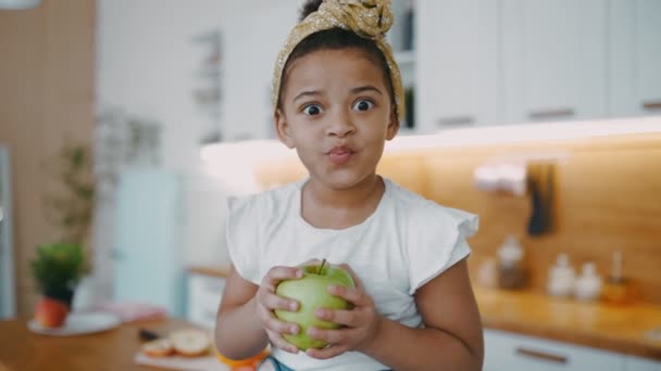 Bella piccola ragazza afro-americana con i capelli ricci siede sul tavolo con mela fresca verde nelle sue mani luce cucina soleggiata in accogliente appartamento — Video Stock