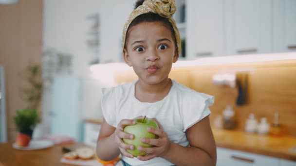 Pretty little african american girl with curly hair sits on table with green fresh apple in her hands light sunny kitchen in cozy apartment — стокове відео