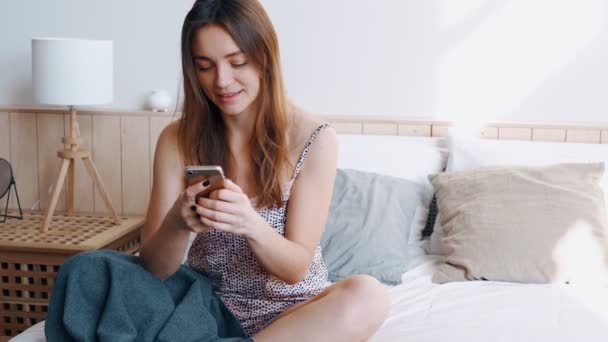 Hermosa chica bastante sonriente con el pelo rubio brillante suelto se sienta en la cama cómoda — Vídeos de Stock