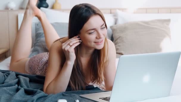 Happy active uśmiechnięta dziewczyna w piżamie rozmawia z przyjaciółmi w sieciach społecznościowych za pomocą słuchawek bezprzewodowych i nowoczesny laptop, młoda piękna kobieta leżąca na miękkim wygodnym łóżku i machając pozdrowienia. — Wideo stockowe