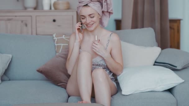 Красивая девушка сидит на диване в квартире собирается на вечеринку, красит ногти с педикюром лаком — стоковое видео