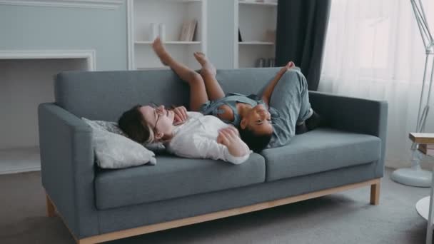 在现代公寓的概念中，年轻纤细和谐的女人在沙发上讨论孩子们一天的问题计划友好的家庭快乐的女儿躺在妈妈的肚子上孩子活泼的孩子和妈妈玩得开心 — 图库视频影像