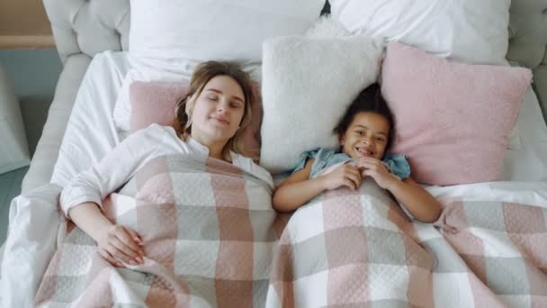 Top-down uitzicht op aantrekkelijke jonge moeder glimlachend liggend op bed met vrolijke Afrikaanse Amerikaanse dochter, portret van mama en zwart meisje liggend onder deken in gezellig appartement overdag slapen — Stockvideo