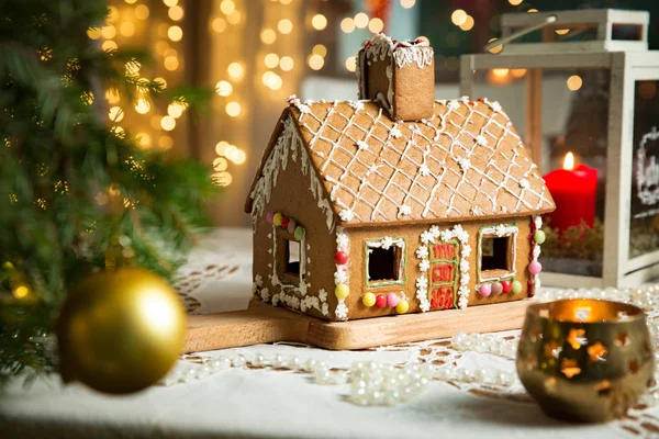 Little gingerbread house with glaze standing on table with tablecloth and decorations, candles and lanterns. Living room with lights and Christmas tree. Holiday mood