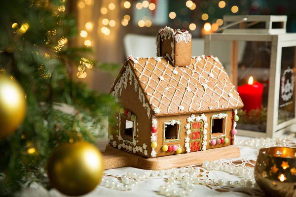 Little gingerbread house with glaze standing on table with tablecloth and decorations, candles and lanterns. Living room with lights and Christmas tree. Holiday mood