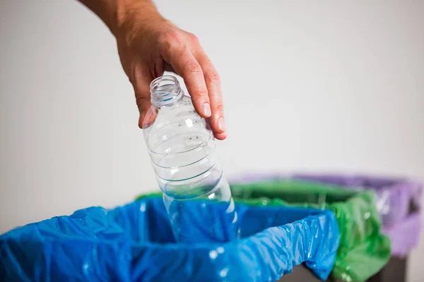 Hand putting single-use plastic bottle into recycling bin. Person in a house kitchen separating waste. Black trash bin with blue bag and recycling symbol.