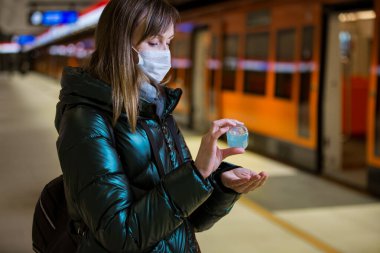 Woman in winter coat with protective mask on face standing on metro station, using hand sanitizer, looking worried. Preventive measures in public places of epidemic regions. Finland, Espoo clipart