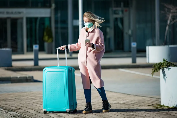 スマートフォンや大きな荷物ケースで空港の入り口に立って保護マスクの女性 ブラウジング テキストメッセージ モバイルアプリを使用して 安全な旅行 — ストック写真