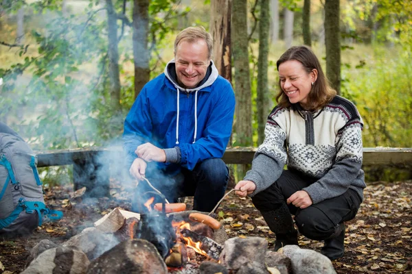 Man and woman roasting on campfire in forest on shore of lake, making a fire, grilling. Happy couple exploring Finland. Scandinavian landscape.