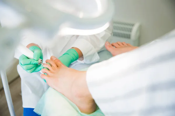 Woman sitting in chair, having foot treatment in spa salon.  Professional medical pedicure specialist in protective mask using special electric lathe and tools. Close-up feet