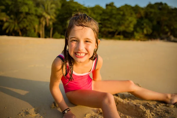 Cute happy little girl playing with sand on the beach in swimming suit, drawing a heart and writing. Beautiful summer sunset, sea, coconut palms, picturesque exotic landscape. Phuket, Thailand
