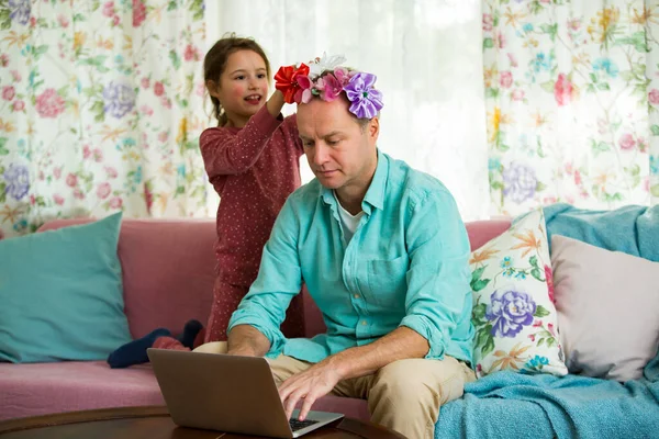 Child playing and disturbing father working remotely from home. Little girl combing daddy\'s hair and making hairstyle. Man sitting on couch with laptop. Family spending time together indoors.